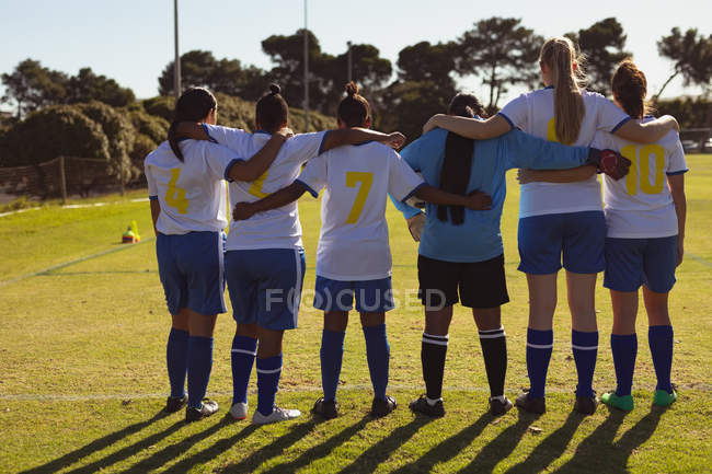 Vista trasera de diversas jugadoras de fútbol de pie con el brazo alrededor en el campo de deportes - foto de stock