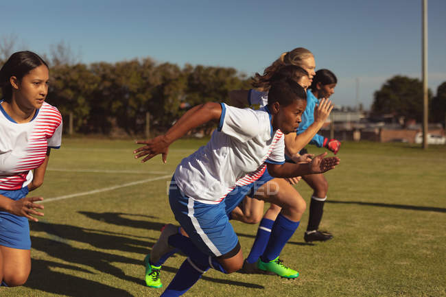 Vista lateral de diversas jugadoras de fútbol corriendo en el campo de deportes en un día soleado - foto de stock