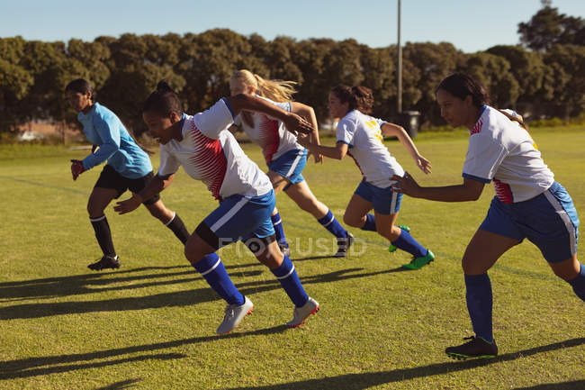 Vista lateral de diversas jugadoras de fútbol corriendo en el campo de deportes en un día soleado - foto de stock