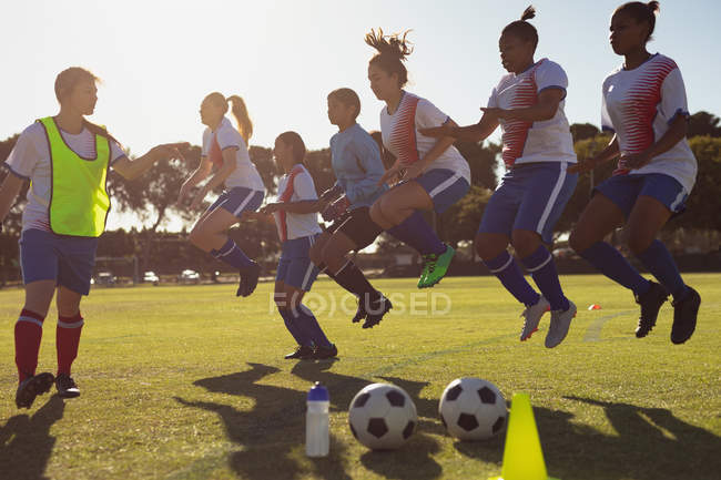Vista lateral del entrenador caucásico ayudando a diversas jugadoras de fútbol con ejercicio de salto en el campo de deportes - foto de stock
