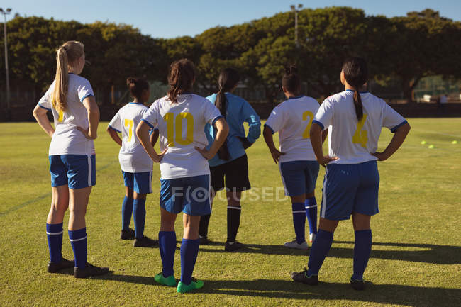 Visão traseira de diversas jogadoras de futebol do sexo feminino de pé com as mãos no quadril no campo esportivo — Fotografia de Stock