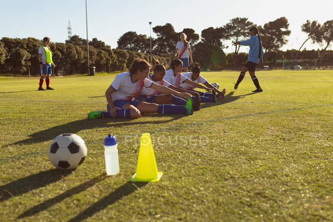 Vista lateral de diversas jugadoras de fútbol haciendo ejercicio de estiramiento en el campo en un día soleado - foto de stock
