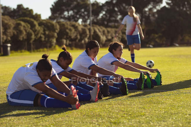 Vista lateral de diversas jogadoras de futebol fazendo exercícios de alongamento em campo em um dia ensolarado — Fotografia de Stock