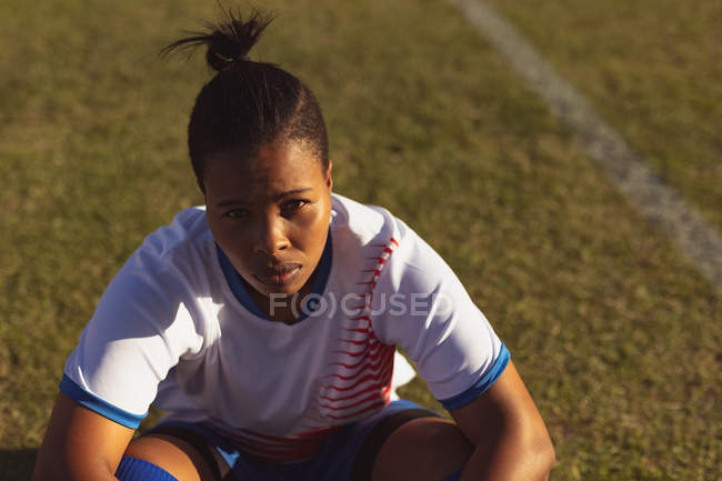 Vista de alto ángulo de la agotada jugadora de fútbol afroamericana sentada en el campo después de un entrenamiento en un día soleado - foto de stock