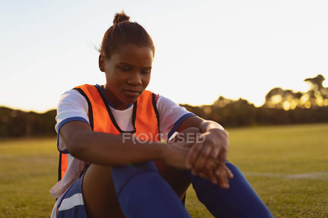 Vista frontal do jogador de futebol feminino afro-americano relaxando na grama no campo esportivo — Fotografia de Stock