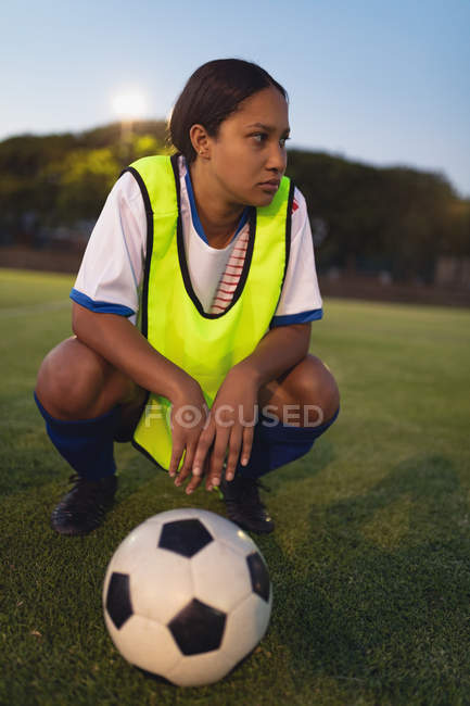Крупный план вдумчивой афроамериканской женщины-футболистки, приседающей с футболом на спортивном поле — стоковое фото