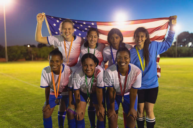 Vista frontal da equipe de futebol feminino diversificada posando com bandeira americana no campo esportivo — Fotografia de Stock