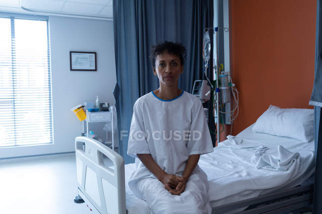 Portrait d'une belle femme métisse d'âge moyen en robe d'hôpital assise sur le lit à l'hôpital — Photo de stock