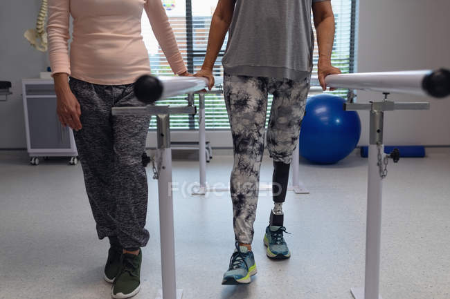 Sección baja del fisioterapeuta femenino que ayuda a la paciente amputada a caminar con barras paralelas en el hospital - foto de stock