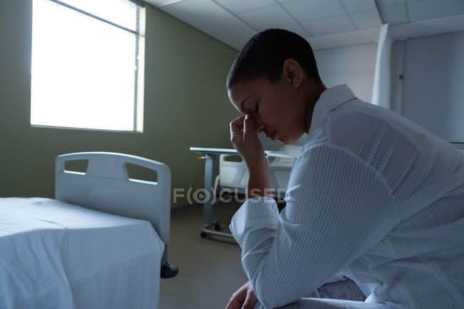 Side view of worried mixed race female patient sitting on bed in the ward in hospital. Empty beds are visible in the background. — Stock Photo