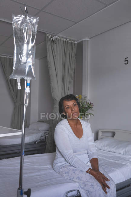 Вид спереди на вдумчивую пациентку смешанной расы, сидящую на кровати с капельницей, стоящей перед ней в палате больницы — стоковое фото