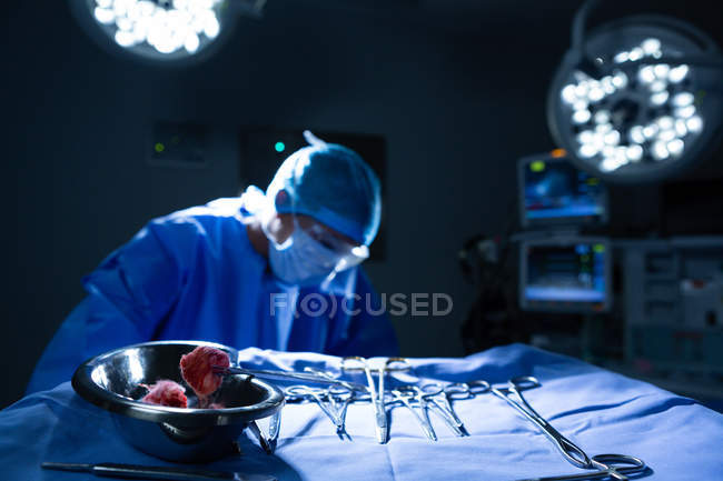 Vista frontal de los instrumentos quirúrgicos y el plato de riñón sobre una mesa, mientras que la joven cirujana de raza mixta trabaja detrás de ella en el quirófano del hospital - foto de stock