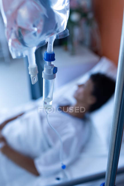 Close-up of intravenous drip with mixed-race female patient lying in bed on the background in the ward at hospital — Stock Photo