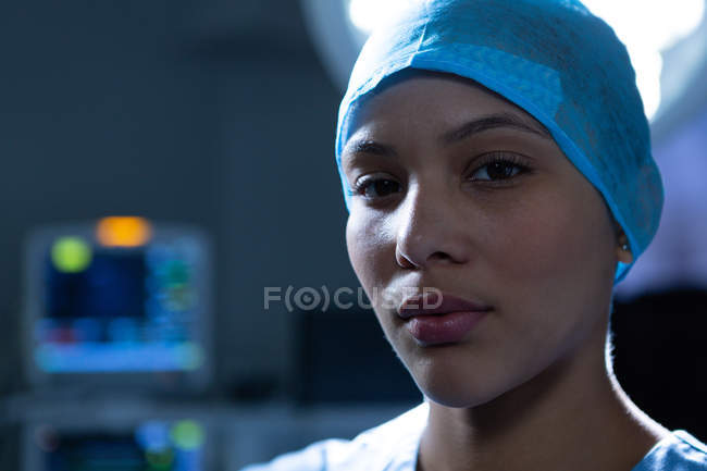 Retrato de una joven cirujana de raza mixta de pie mientras mira la cámara en la sala de operaciones del hospital. Cirujano lleva gorra quirúrgica y bata . - foto de stock