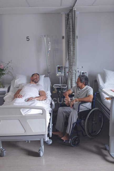 Vista laterale di diversi pazienti maschi che interagiscono tra loro nel reparto ospedaliero. Paziente maschio caucasico sdraiato a letto mentre il paziente di razza mista si siede sulla sedia a rotelle . — Foto stock