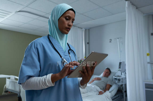 Visão de baixo ângulo da bela mestiça médica com hijab usando comprimido digital na enfermaria enquanto o paciente masculino caucasiano dorme em segundo plano no hospital — Fotografia de Stock