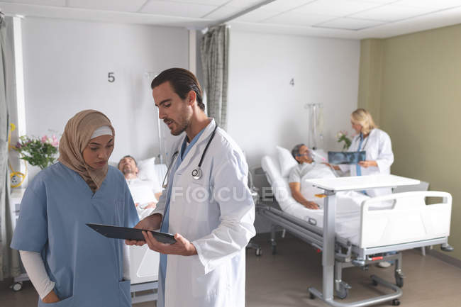 Vue de face de divers médecins discutant sur presse-papiers dans le service de l'hôpital. En arrière-plan, une femme médecin caucasienne parle de rayons X avec un patient masculin de race mixte âgé . — Photo de stock