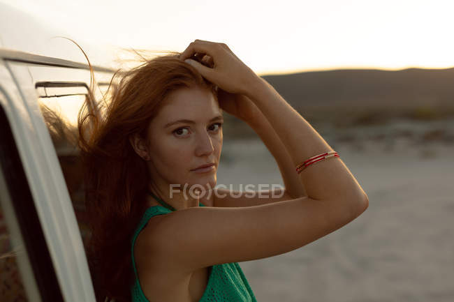 Side view of beautiful Caucasian woman looking at camera while leaning on camper van at beach — Stock Photo