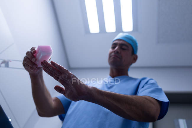 Низький кут зору красивий середній вік Кавказького чоловічого хірурга очищення рук з щіткою і милом в раковину в лікарні. Він носить хірургічне плаття і шапочка. — стокове фото