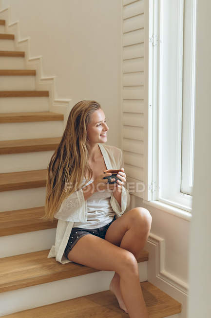Вид збоку щаслива кавказька жінка з чашкою кави переглядаючи вікно, сидячи на сходах в домашніх умовах — стокове фото