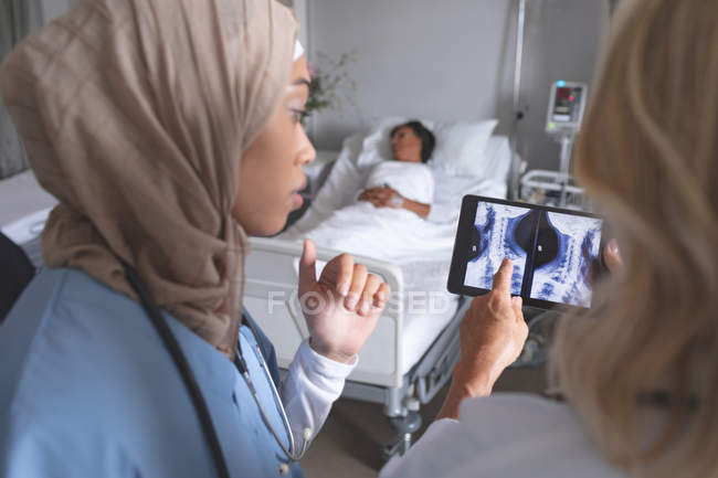 Primer plano de diversas doctoras discutiendo sobre el informe de rayos X en tabletas digitales en la sala del hospital. En el fondo, la paciente de raza mixta está durmiendo en la cama en la sala del hospital. . - foto de stock