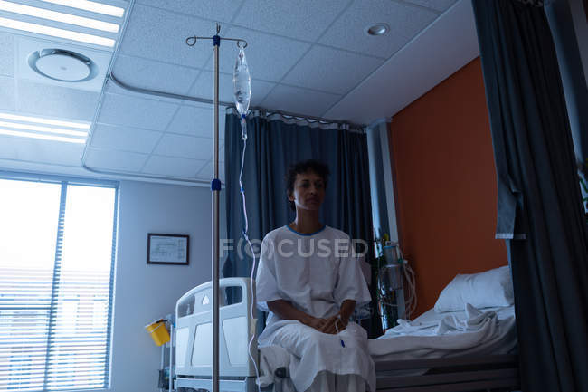 Vorderansicht einer nachdenklichen Patientin mittleren Alters mit gemischter Rasse, die auf dem Bett sitzt, während sie im Krankenhaus behandelt wird — Stockfoto