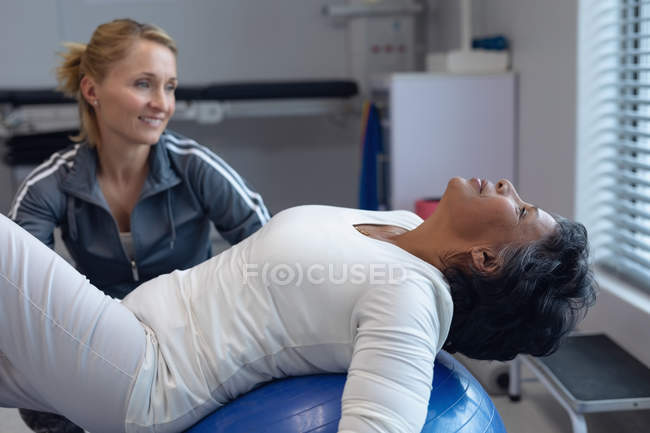 Vista lateral de fisioterapeuta femenina caucásica dando fisioterapia a una paciente de raza mixta en una pelota de ejercicio en el hospital - foto de stock