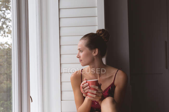 Вид спереди кавказской женщины с кофейной кружкой, смотрящей в окно дома — стоковое фото