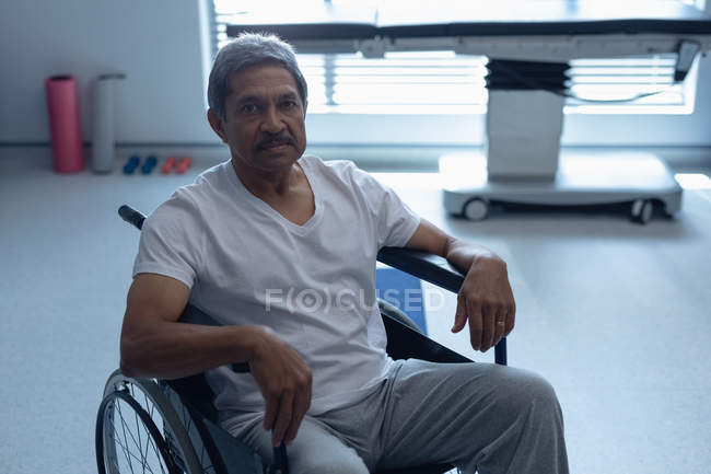Vista frontal del paciente masculino de raza mixta mirando la cámara mientras está sentado en silla de ruedas en el hospital - foto de stock