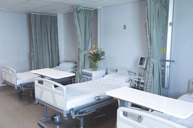 Modern hospital ward with empty beds, medical monitor, green curtains, cupboards, and flowers. — Stock Photo