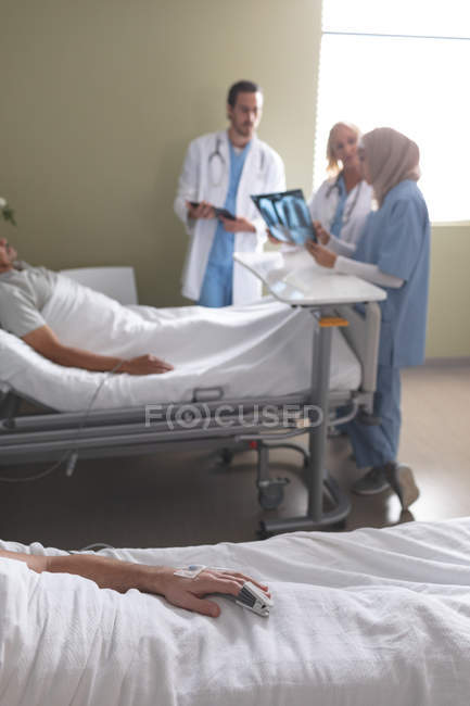 Seitenansicht eines kaukasischen männlichen Patienten, der es sich auf dem Bett bequem macht, während verschiedene Ärzte auf der Station im Krankenhaus miteinander interagieren. im Vordergrund schläft ein kaukasischer Patient im Bett. — Stockfoto