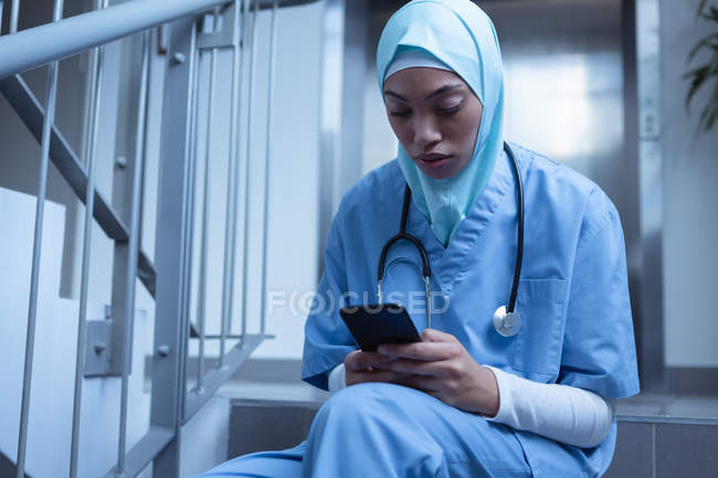 Vista frontal da enfermeira mestiça no hijab usando telefone celular nas escadas do hospital — Fotografia de Stock