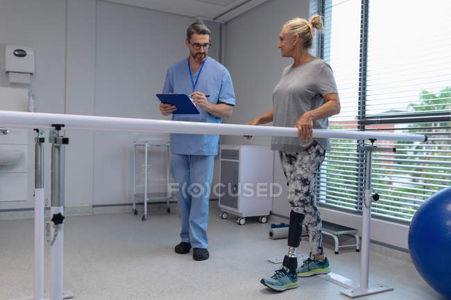 Vista frontal de la escritura del fisioterapeuta masculino caucásico en el portapapeles y paciente amputado femenino caucásico caminando con barras paralelas en el hospital - foto de stock