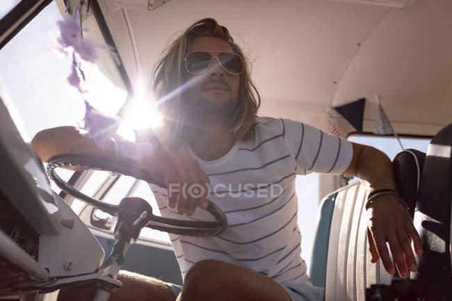 Baixo ângulo de visão de belo jovem caucasiano com óculos de sol olhando para longe na van na praia — Fotografia de Stock