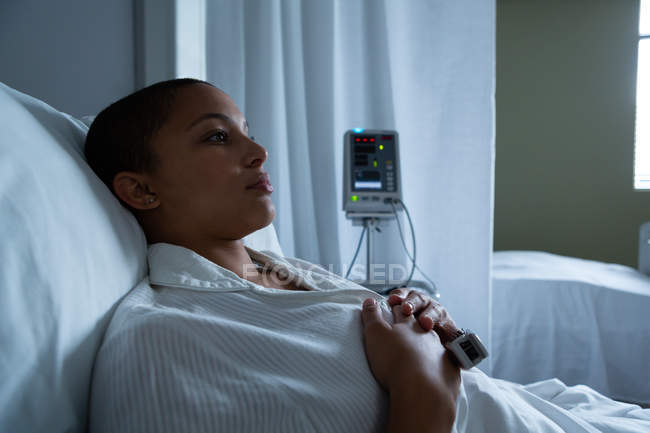 Side view of thoughtful mixed race female patient lying in bed with hands on her breasts in the ward in hospital. Empty bed and monitor are visible in the background. — Stock Photo
