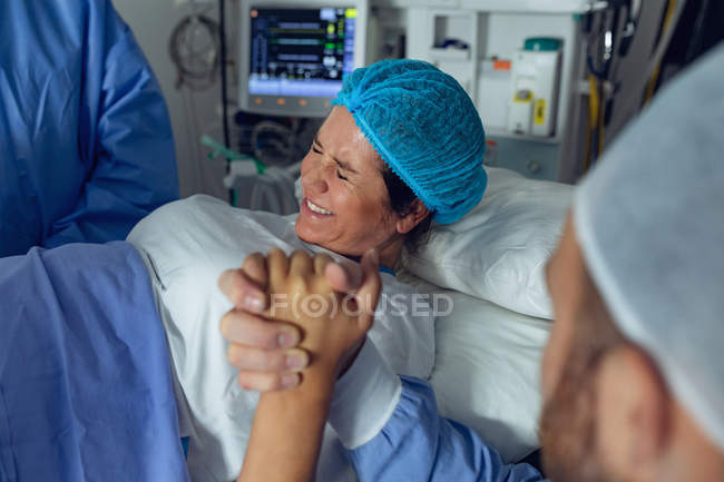 Seitenansicht des Chirurgen, der schwangere Frau während der Entbindung untersucht, während Mann ihre Hand im Operationssaal im Krankenhaus hält — Stockfoto
