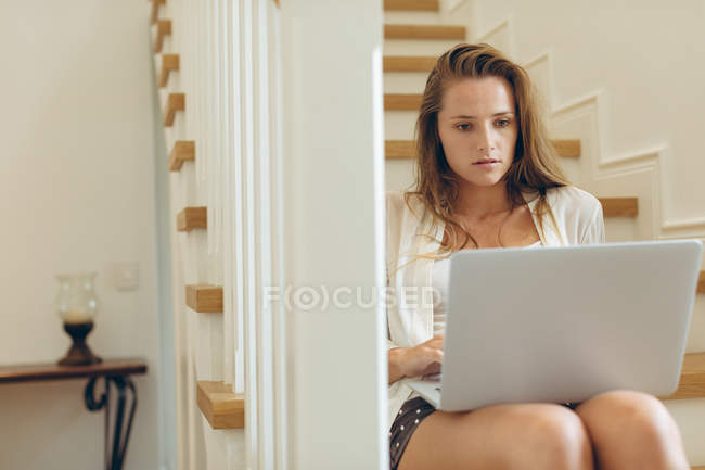 Vista frontal de la hermosa mujer caucásica utilizando el ordenador portátil en las escaleras en casa - foto de stock