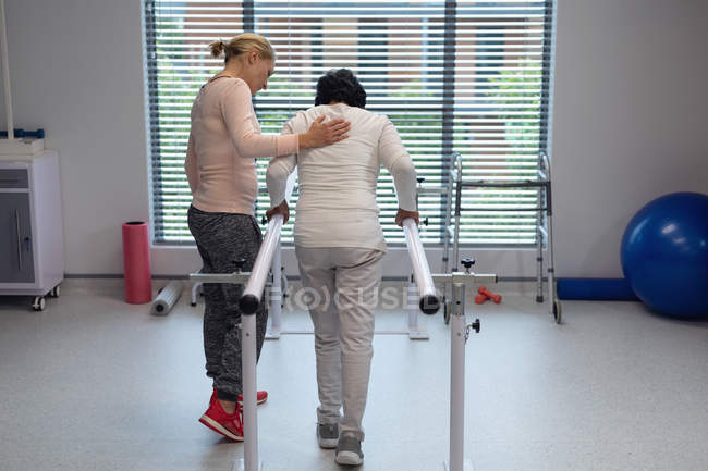 Rear view of Caucasian female physiotherapist helping mixed-race female patient walk with parallel bars in the hospital — Stock Photo