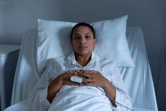 Portrait of beautiful young mixed race female patient lying on bed with hands on stomach in the ward in hospital. Pulse oximetry is measuring the oxygen level in her blood. — Stock Photo