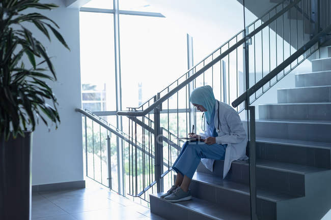 Vista lateral de la doctora de raza mixta en hiyab escribiendo en el portapapeles en la escalera en el hospital - foto de stock
