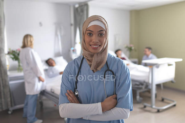 Ritratto di dottoressa mista in hijab in piedi con le braccia incrociate in ospedale. Sullo sfondo diversi medici stanno interagendo con i loro pazienti
. — Foto stock