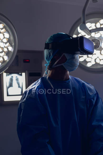 Vorderansicht einer kaukasischen Chirurgin mit Virtual-Reality-Headset im Operationssaal des Krankenhauses. Im Hintergrund sind medizinisches Licht und Röntgenbilder zu sehen. — Stockfoto