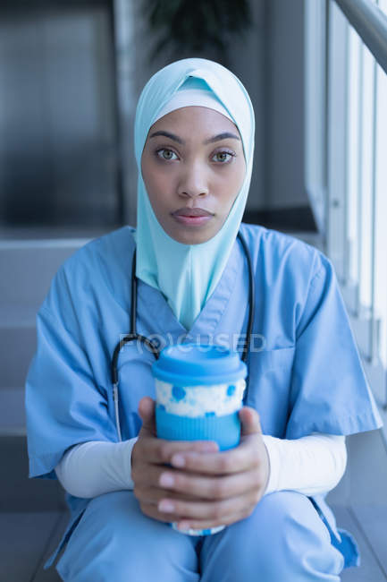 Portrait of mixed race female doctor in hijab having coffee on staircase in hospital — Stock Photo