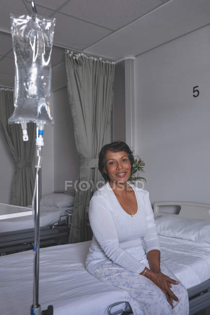 Retrato de una hermosa paciente de raza mixta sentada en la cama mientras sonríe hacia la cámara en la sala del hospital. IV stand está al lado de la cama . - foto de stock