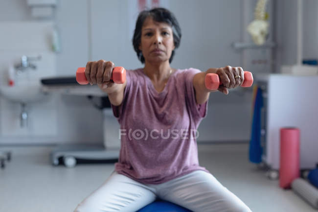 Vista frontal de una paciente de raza mixta que hace ejercicio con pesas sobre una pelota de ejercicio en el hospital - foto de stock