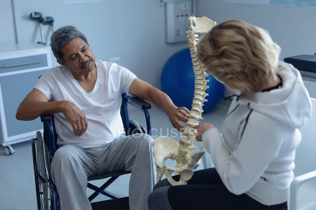 Vista posteriore del fisioterapista femminile caucasico che spiega il modello della colonna vertebrale al maschio di razza mista in sedia a rotelle in ospedale — Foto stock