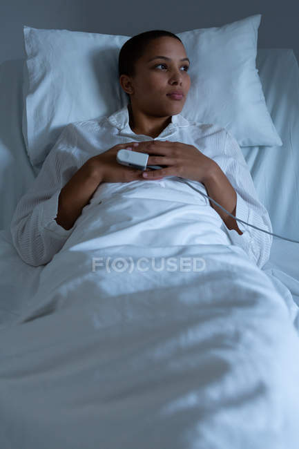 Front view of beautiful thoughtful mixed race female patient lying on bed in the ward while looking outside in hospital. Pulse oximetry is measuring measuring the oxygen levels in her blood. — Stock Photo