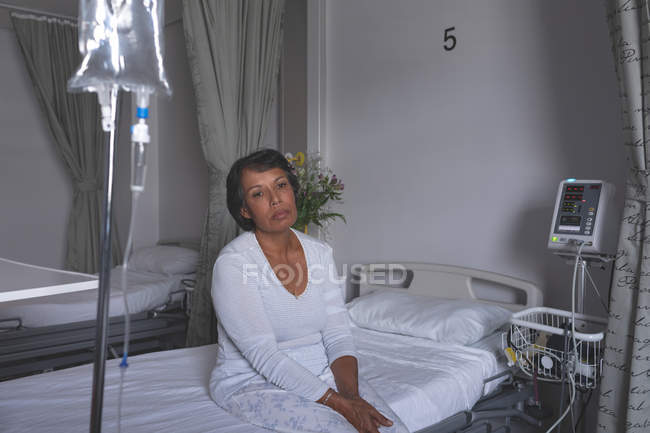 Front view of thoughtful mature mixed-race female patient sitting on bed in the ward at hospital — Stock Photo
