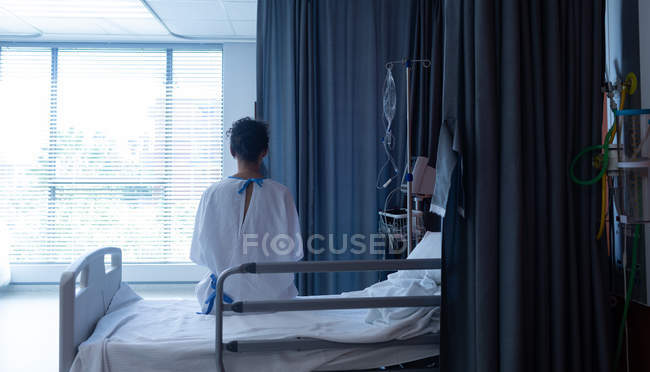 Rear view of thoughtful mixed-race female patient in hospital gown sitting on the bed while looking outside in hospital — Stock Photo