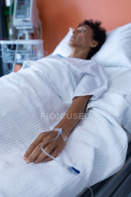Front view of thoughtful mixed-race female patient lying on bed while getting IV therapy in the ward at hospital — Stock Photo